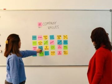 two women standing in front of a white board with sticky notes on it