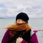 A person with long red hair blowing across their face on a beach