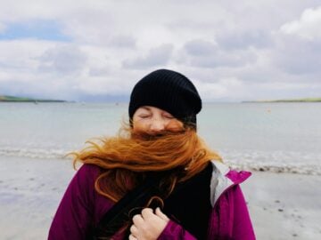 A person with long red hair blowing across their face on a beach