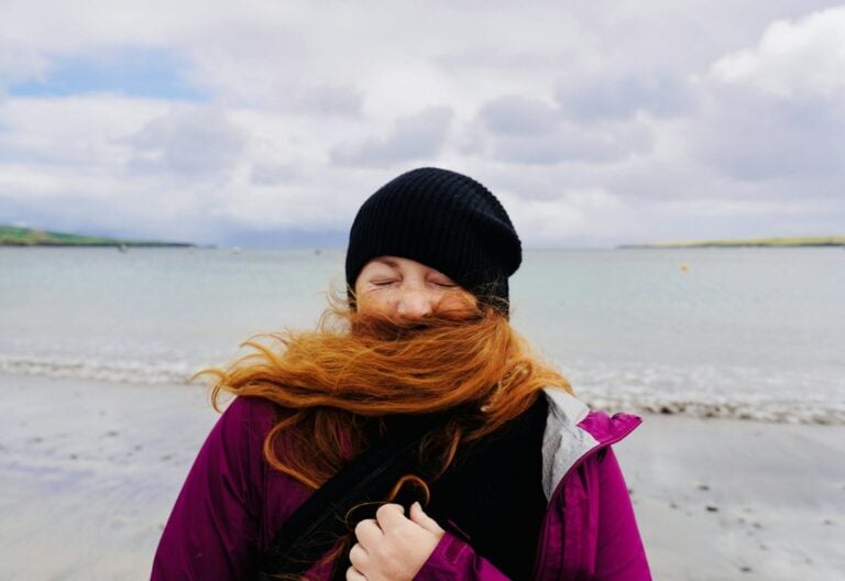 A person with long red hair blowing across their face on a beach